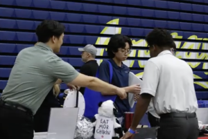 A recruiter gestures while speaking with two students at a job fair booth, which features a plush cow toy wearing a sign that reads 'EAT MOR CHIKIN.' inside College of the Canyons gymnasium.